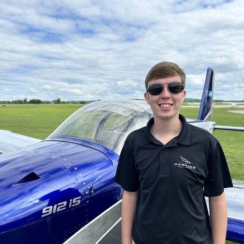 Caleb Kahler in sunglasses and a Hawkins Flight Academy polo standing beside the canopy of a blue RV-12 with green fields behind