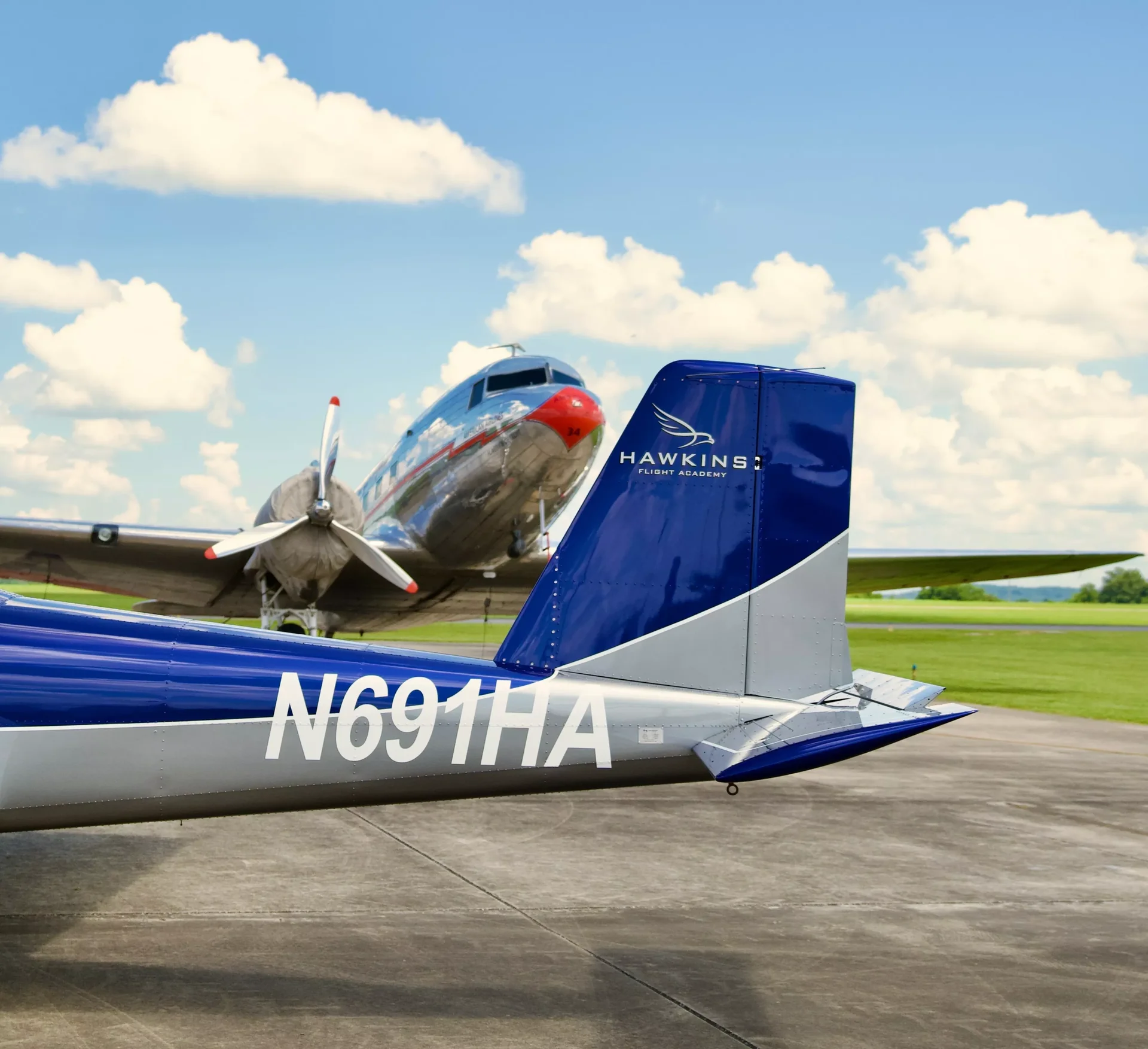 Hawkins-branded RV-12 tail in the foreground with a vintage twin-engine airplane behind it on the ramp