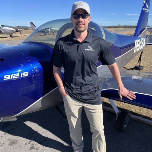 Ian Condon in a Hawkins Flight Academy polo and cap standing in front of a blue RV-12 on a sunny ramp
